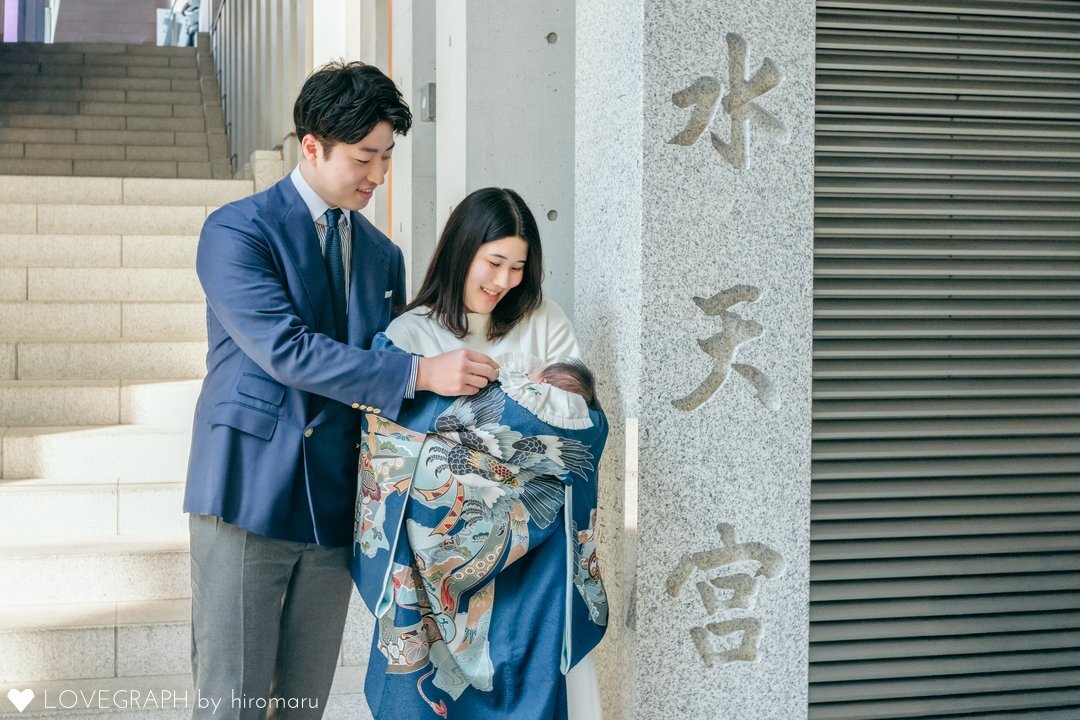 水天宮でお宮参りをしている両親の写真。赤ちゃんは男の子向けの産着、父親は紺のジャケット、母親は白いワンピースを着用している。