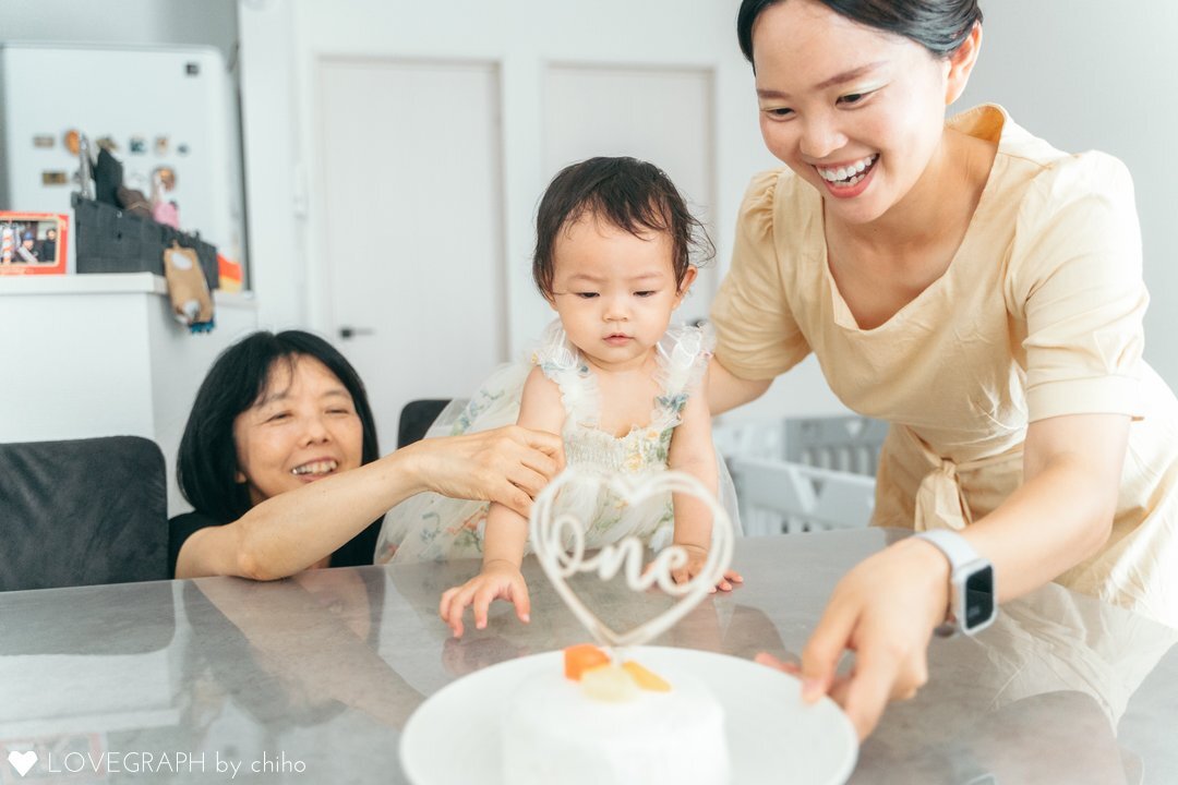 ケーキを警戒する赤ちゃんの手をとって、ケーキを触らせようとする母親と祖母の写真。緊張感のある赤ちゃんと、反応を楽しむ親の表情が対照的でユーモラス。