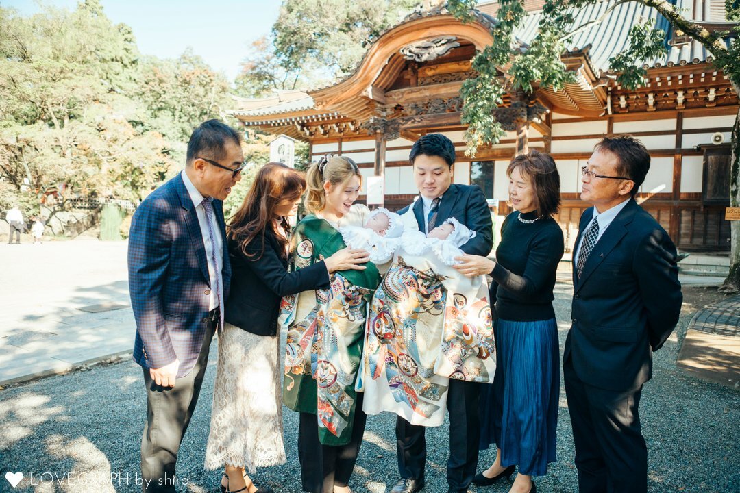 東京都調布市の深大寺でお宮参りを行うご家族の様子。双子の赤ちゃんを中心に、祖父母や両親、叔母（伯母）が並び、国宝釈迦如来像が安置される釈迦堂を背景に、赤ちゃんを見つめ合う温かい記念写真。