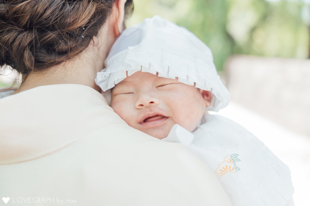 お宮参りで着物姿の母親の肩に抱かれ、穏やかな寝顔を見せる赤ちゃんのアップ写真。背景の緑が優しくボケる自然光の中、赤ちゃんの白い産着や帽子が映える温かいポートレート。場所は東京都港区の増上寺