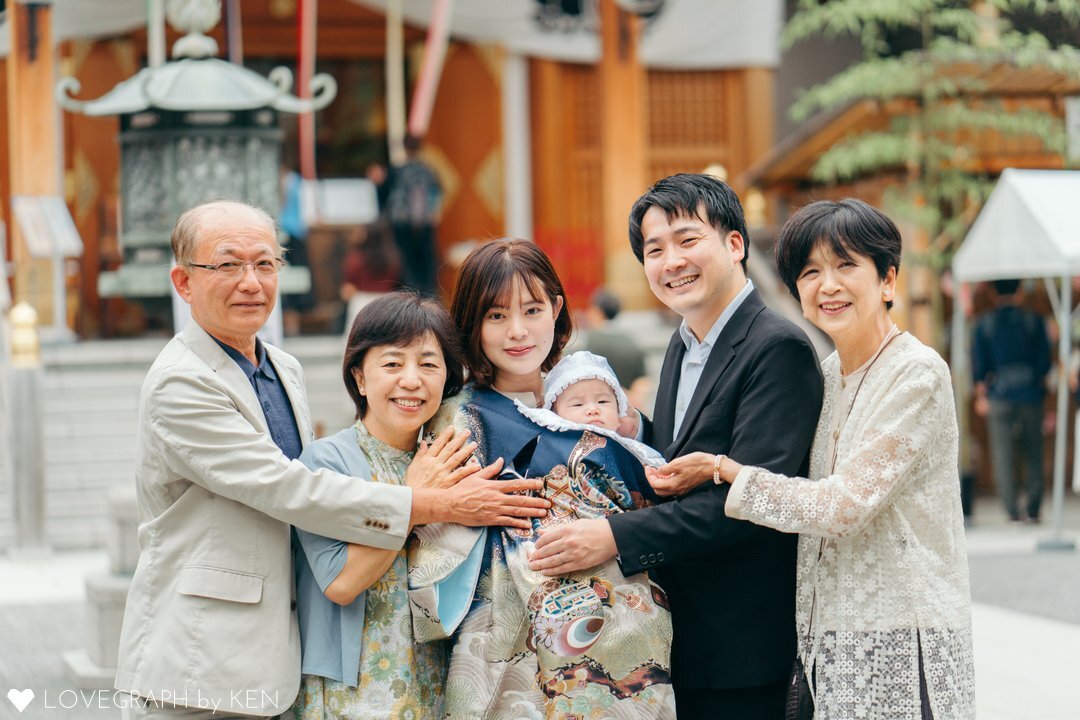 東京・日本橋の水天宮でお宮参りを行うご家族の様子。荘厳な社殿を背景に、赤ちゃんを抱く母親と寄り添う父親、そして祖父母が並び、紺色の豪華な祝い着（産着）を掛けた赤ちゃんを優しく見つめる、幸せあふれる三世代の記念写真。