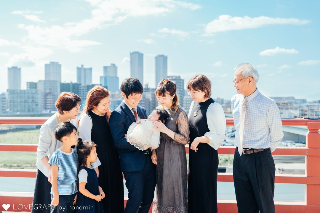 東京都大田区の多摩川浅間神社にある「見晴台」でお宮参りを行うご家族の様子。赤ちゃんを中心に、祖父母、両親、上の子2人が並び、多摩川と武蔵小杉のスカイラインを背景に、晴天の下、赤ちゃんを見つめ合う温かい記念写真。