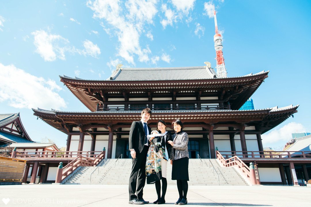 東京の増上寺でお宮参りを行うご家族の様子。荘厳な本堂を背景に、赤ちゃんを抱く母親、父親、そしておばあちゃん（祖母）が並び、その背後には東京タワーがそびえ立つ、伝統と現代が融合した記念写真。