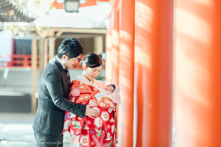 東京の赤坂にある山王日枝神社の「千本鳥居」でお宮参りの記念撮影をする家族。鮮やかな赤い祝い着（産着）を掛けた赤ちゃんを抱く着物姿の母親と、寄り添うスーツ姿の父親が、並ぶ赤い鳥居の光の中で微笑む温かい写真。
