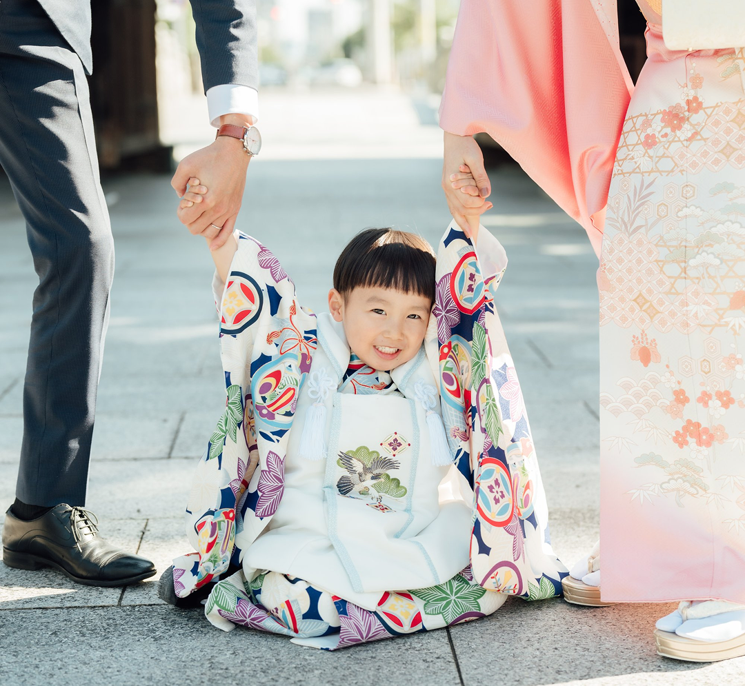 七五三の記念写真：白い被布（ひふ）を着用して参道に座り込み、両親と手を繋いで微笑む3歳の男の子