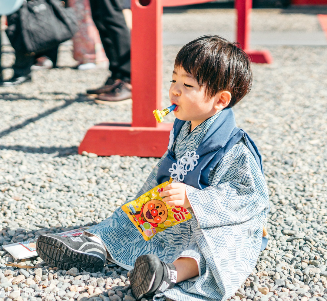 七五三の風景。青い着物姿の3歳の男の子が地面に座り込み、お菓子（グミ）を持っている。口には笛を咥えている。