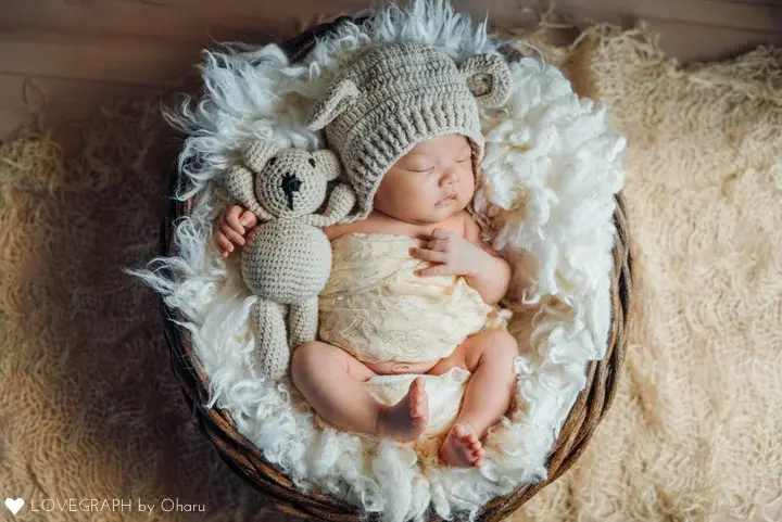 アートニューボーンの作例。籐のバスケットの中で白いファーに包まれて眠る新生児。クマの耳がついたニット帽をかぶり、隣にはお揃いの編みぐるみのクマが置かれている。