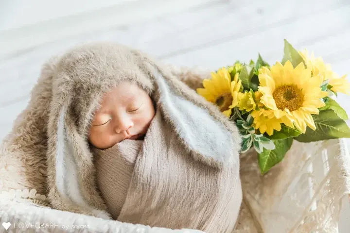 アートニューボーンの作例。ベージュのふわふわしたウサギのコスチュームでおくるみされ、ぐっすり眠る赤ちゃんのアップ。横には鮮やかな黄色のひまわりが添えられている。