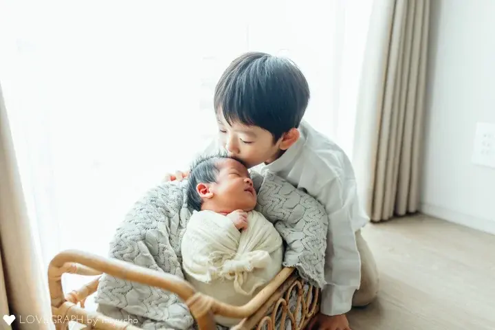 ナチュラルニューボーンの作例。籐のベビーベッドでおくるみに包まれた新生児と、その赤ちゃんの額に優しくキスをする兄の姿。窓からの自然光に包まれた温かい雰囲気の兄弟写真。