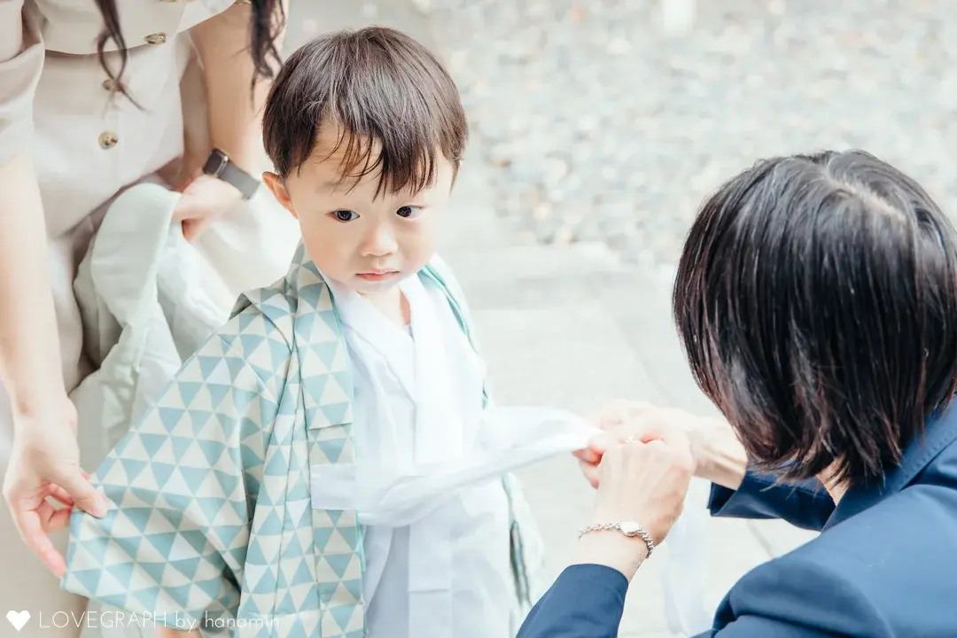 水色と白の鱗文様の羽織を身にまとい、着付けをしてもらう小さな男の子。周囲の大人の手が、男の子の着物や帯を丁寧に整えている様子。