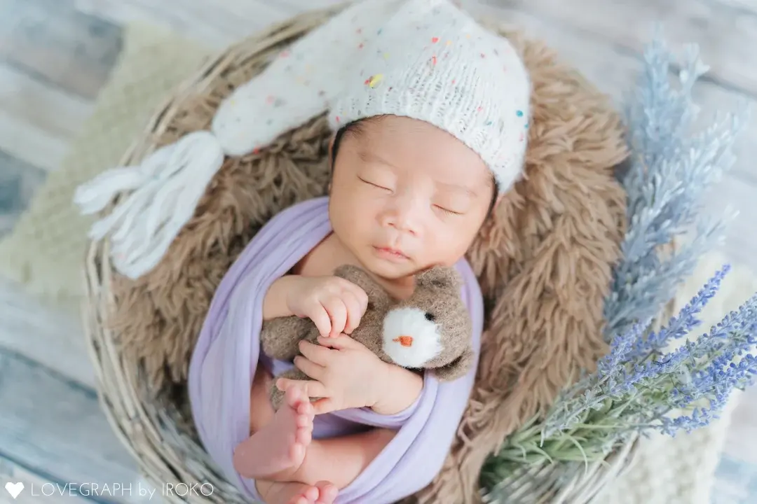 カゴの中でクマのぬいぐるみを抱いて眠る赤ちゃんのニューボーンフォト。ラベンダー色のおくるみに包まれ、カラフルなドットが入った白いニット帽を被っています。周りにはラベンダーなどの草花が添えられ、全体的に明るくソフトなトーンでまとめられています。