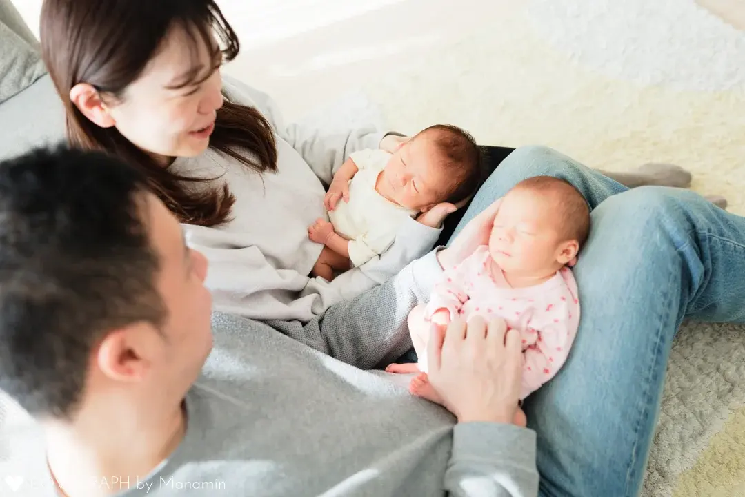 パパとママの膝の上で、それぞれ一人ずつ抱っこされて眠る双子の赤ちゃんの写真。ご両親は赤ちゃんを優しく支えながら、互いに顔を見合わせて微笑んでいます。自然光が差し込む明るい室内での、温かな家族のひとときを捉えています。