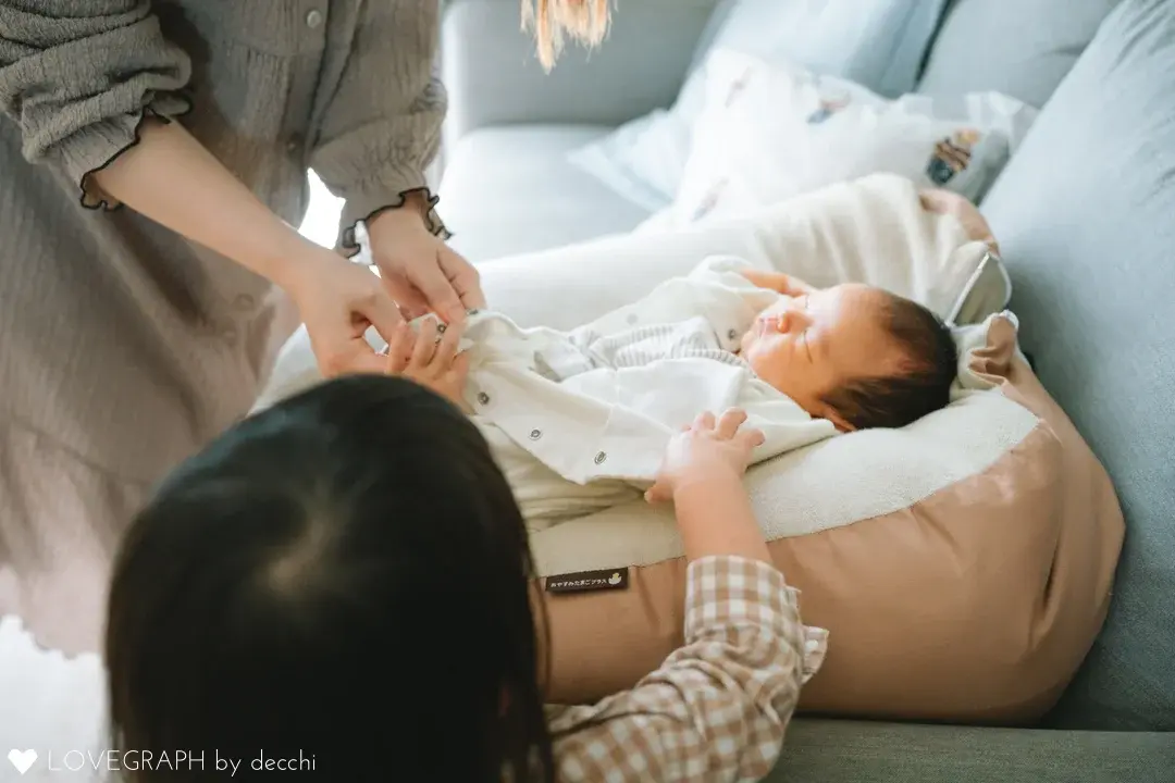 ソファの上のクッションで眠る赤ちゃん。母親が服のボタンを留め、手前では上の子が赤ちゃんにそっと手を伸ばしている様子。