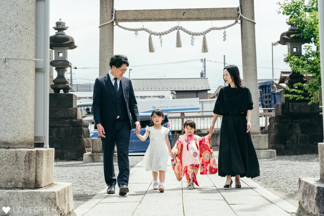 神社の鳥居の前で、手をつないで仲良く歩く4人家族。着物を着た3歳の女の子、白いドレスを着たお姉ちゃん、スーツ姿の父親、黒いワンピースを着た母親の七五三ファミリーフォト。