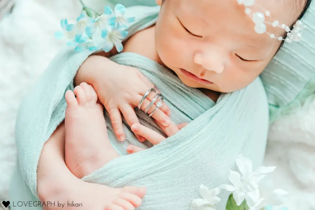 薄いミントグリーンの布に包まれ、気持ちよさそうに眠る新生児。小さな手に両親の結婚指輪をはめ、隣に赤ちゃんの小さな足が見える、愛情に満ちたニューボーンフォト。