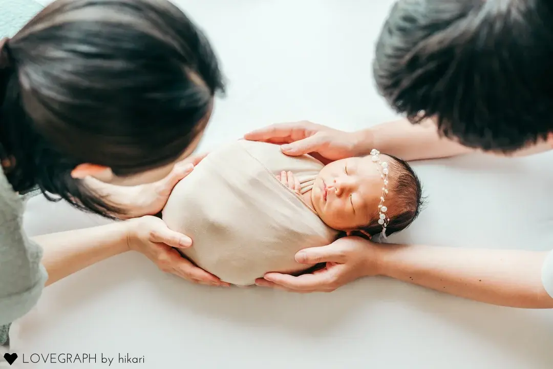 ベージュのおくるみに包まれて眠る新生児を、左右から両親が優しい手つきで支え、愛おしそうに見つめる姿を真上から捉えたニューボーンフォト。家族3人の絆が伝わる温かい1枚。