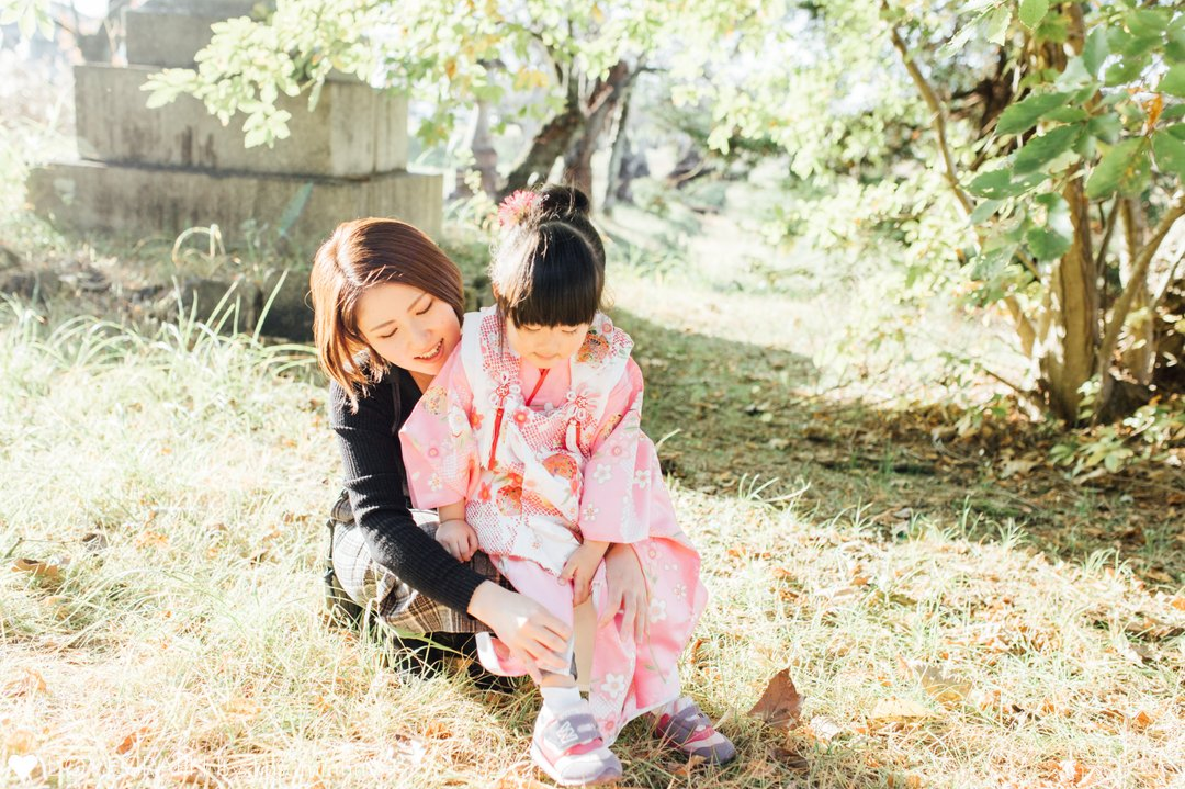 木漏れ日が差す自然の中で、着物姿の女の子の足元に寄り添い、優しくスニーカーを履かせるお母さん。七五三の撮影の合間の、和やかなオフショット写真。