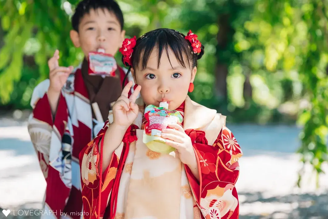 七五三撮影の合間にゼリー飲料で水分補給をする兄妹。カメラに向かって無邪気にピースをする女の子と、後ろで見守るようにお揃いでゼリーを飲む男の子の自然体な一コマ。