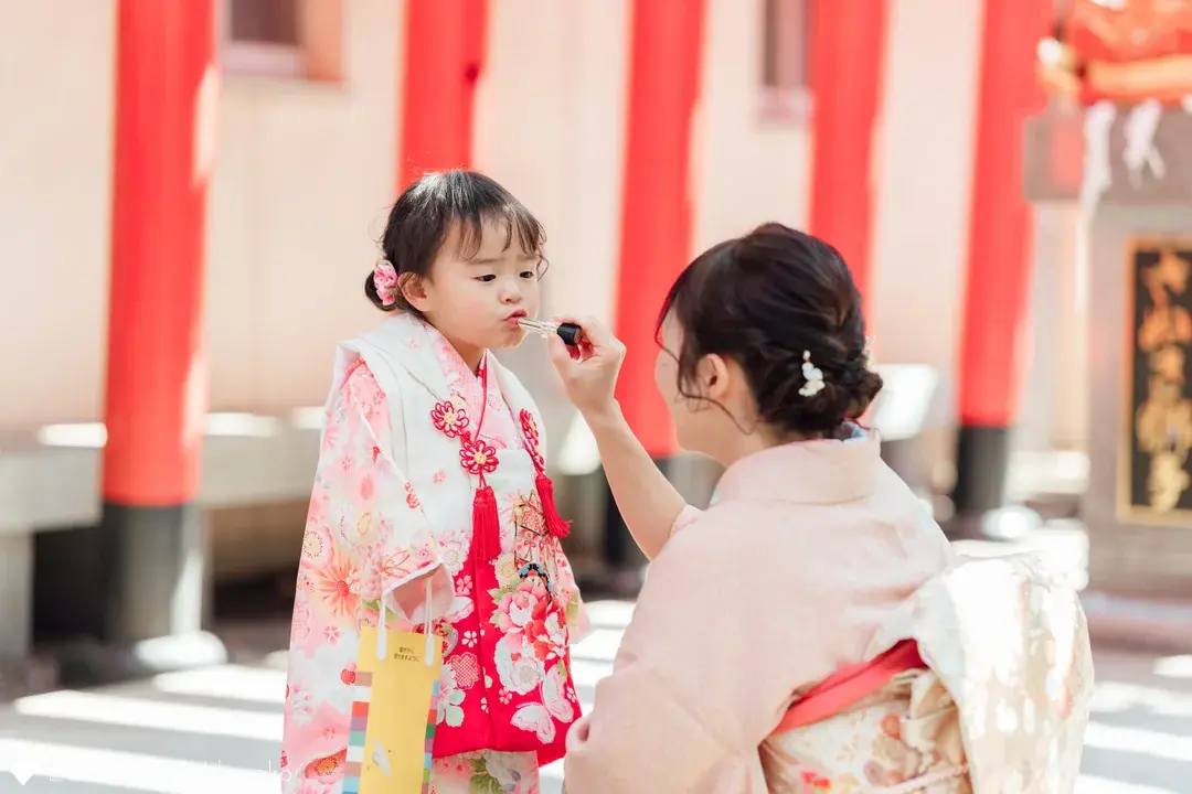 七五三のお参り当日、境内で母親に優しく口紅を塗ってもらう女の子。少し緊張した面持ちで唇を差し出す姿と、着物姿で見守る母親の愛情が伝わるエモーショナルな出張撮影の一コマ。