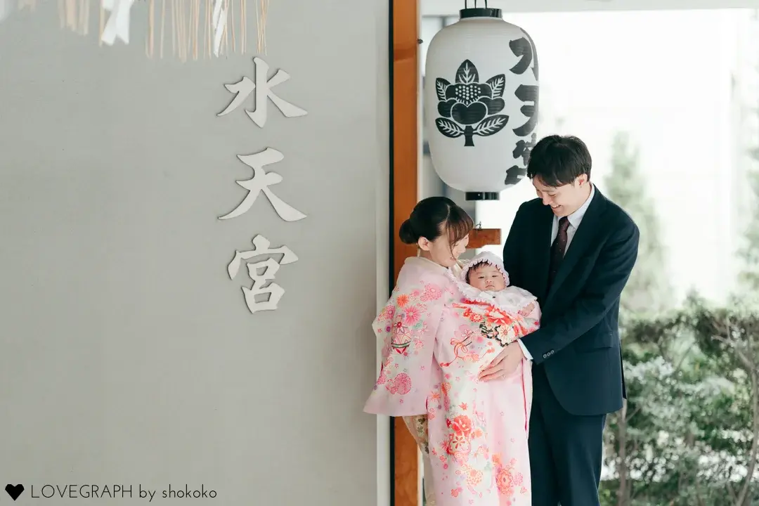 水天宮の文字と提灯の前で、ピンク色の華やかな産着（ピンク地に手鞠と花かご）に包まれた赤ちゃんを抱き、笑顔で見つめ合う両親のお宮参りの写真。