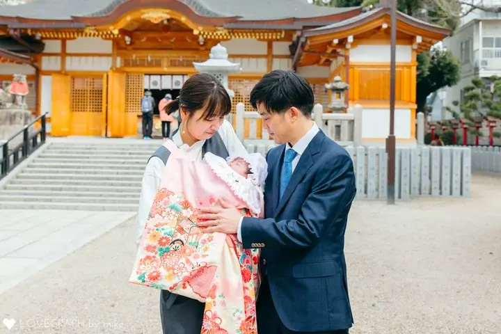 ラブグラフの記事に掲載された、神社での出張撮影例。「ピンククリーム地 季節の花々と鞠の産着」を着用した赤ちゃん、その頭に手を添える父親、そして赤ちゃんを見つめる母親の3ショット。背景には伝統的な拝殿と石畳。