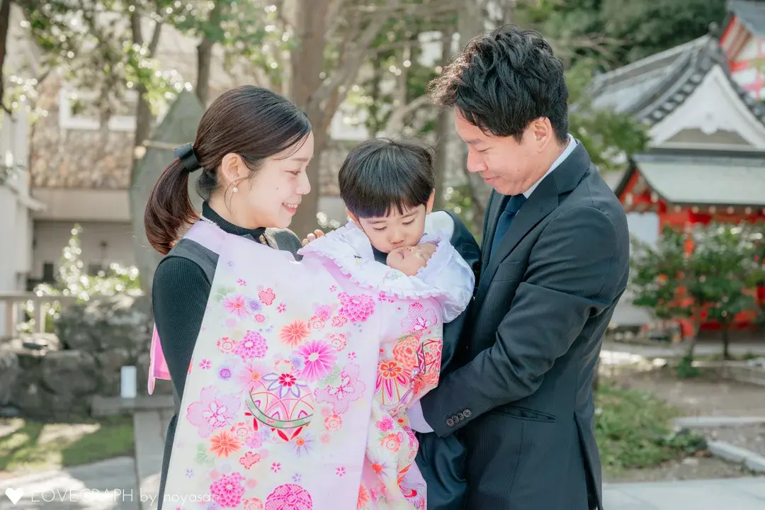 「ピンク地に手毬と花尽くし」産着の着用画像。お宮参りの家族写真。ママに抱っこされピンクの産着に包まれた赤ちゃんに、パパに抱きかかえられたお兄ちゃんがキスをしている。両親は笑顔でその様子を見守っている。