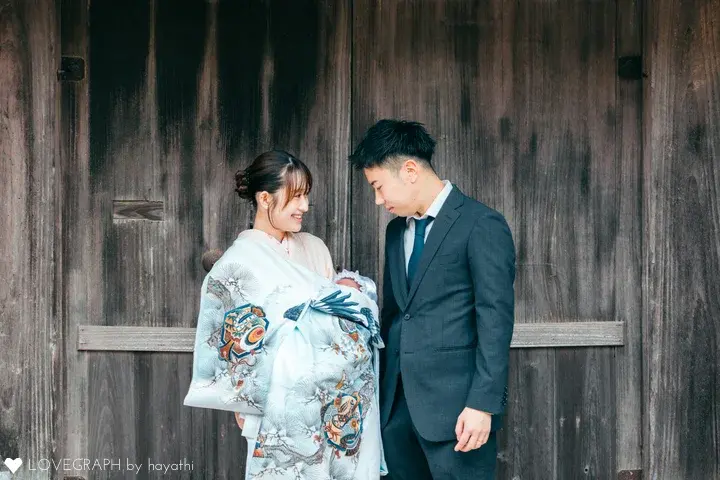 産着「白地に鷹と兜 松と軍配の吉祥柄」の着用写真。神社でのお宮参り。縁起の良い柄が描かれた水色の祝い着の赤ちゃんを抱っこするママと、それを見守るパパが優しく見つめ合う、家族の温かさが伝わる一枚。