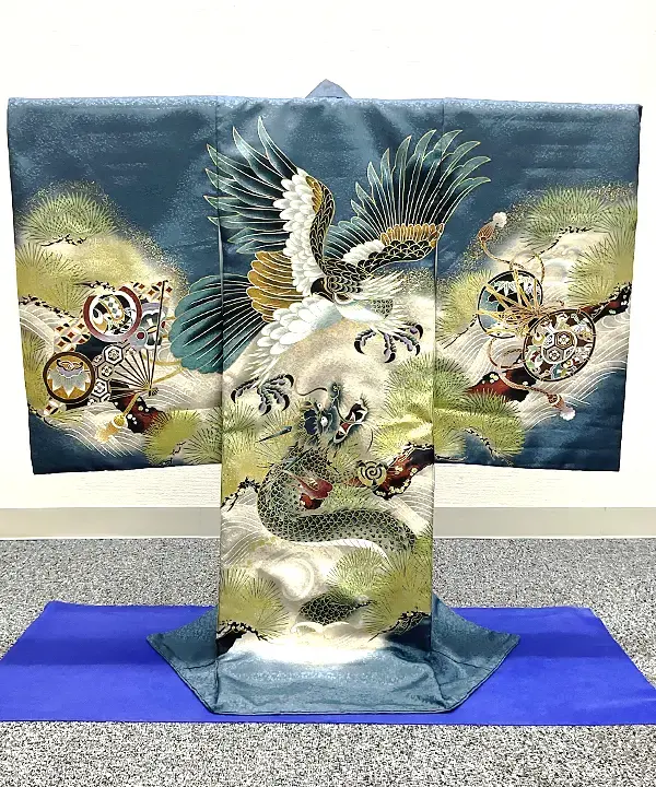 男児用産着「鷹と龍に末広と小槌」の全体画像。深みのある青緑色の生地をベースに、中央には大きく羽を広げた鷹と上を見上げる力強い龍がダイナミックに描かれています。両袖には打ち出の小槌や末広（扇）、背景には縁起の良い松が金彩を交えて華やかにあしらわれています。
