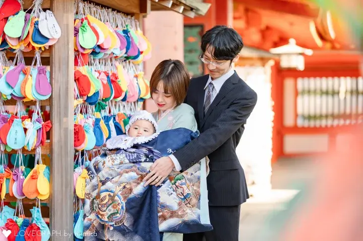 色鮮やかな飾りが掛けられたお堂を背景に、産着「紺色地に鷹と大兜」に包まれた赤ちゃんを抱っこする着物姿の母親と、手を添えて優しく見守るスーツ姿の父親の写真。