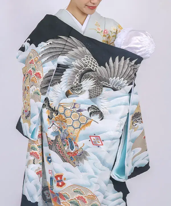産着「鷹と宝船 宝尽くし」の全体画像。鷹や宝船など縁起の良い柄が大きく描かれた男の子用の産着。淡い色の着物を着た女性が、白い帽子をかぶった赤ちゃんを優しく抱っこしている。