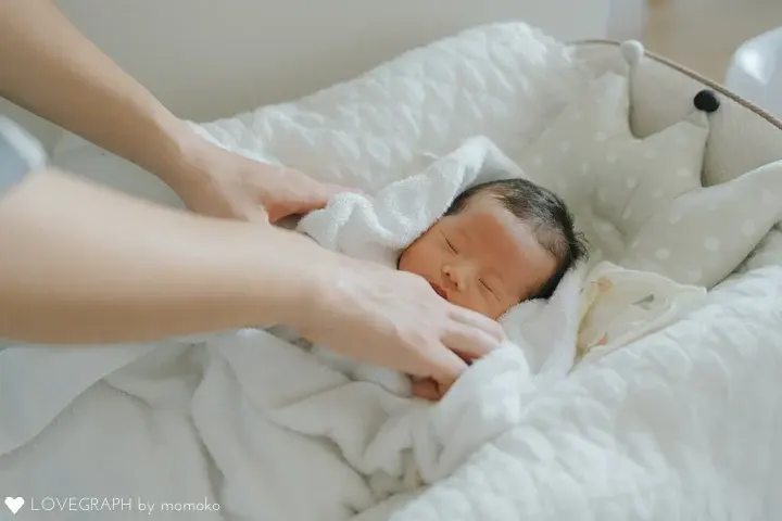 王冠型のクッションの横でスヤスヤと眠る新生児と、赤ちゃんを優しく白いタオルで包み込む親の手