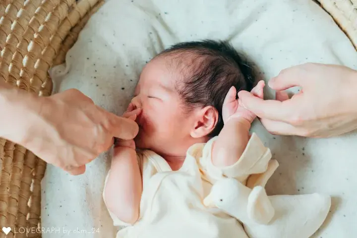 ラブグラフのナチュラルニューボーンフォト。かごのクーファンで安心して眠る赤ちゃんと、パパとママの指をぎゅっと握る温かい触れ合いの様子