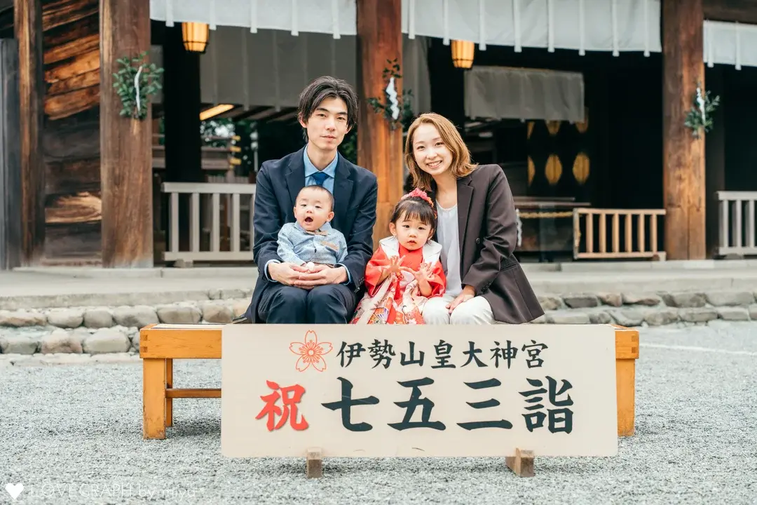 『伊勢山皇大神宮 祝 七五三詣』と書かれた立て看板の後ろで、ベンチに座って記念撮影をする4人家族。スーツ姿のお父さんに抱っこされた赤ちゃん、赤い着物を着て両手を上げる女の子、ジャケット姿のお母さんが並んでカメラを見つめている
