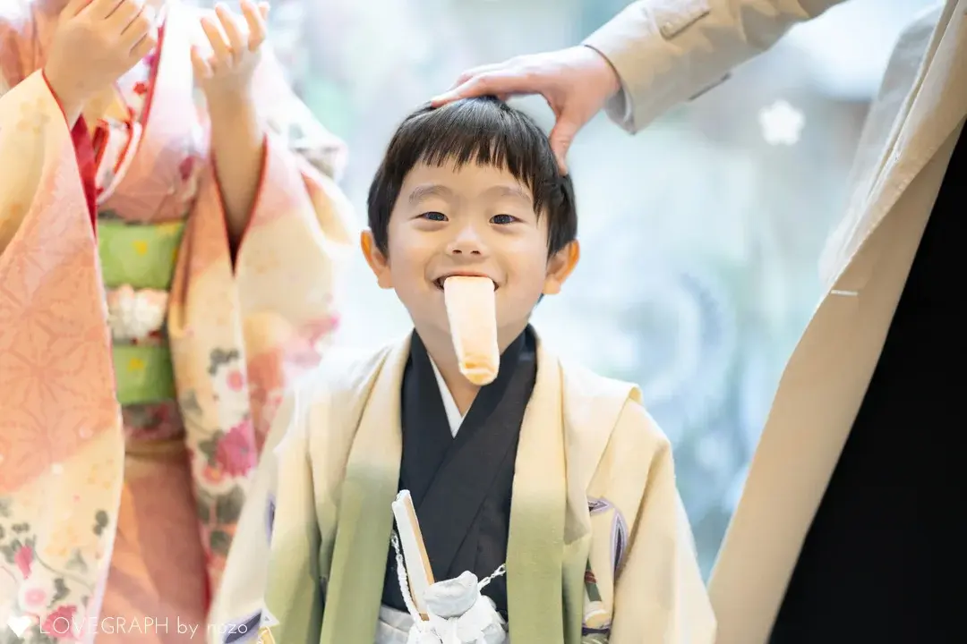 伊勢山皇大神宮の記念館にて七五三の羽織袴を着て、口にスティックパンをくわえながら満面の笑みを浮かべる男の子。隣に立つ父親に頭を優しく撫でられており、左側にはピンク色の着物を着た母親が写っている。