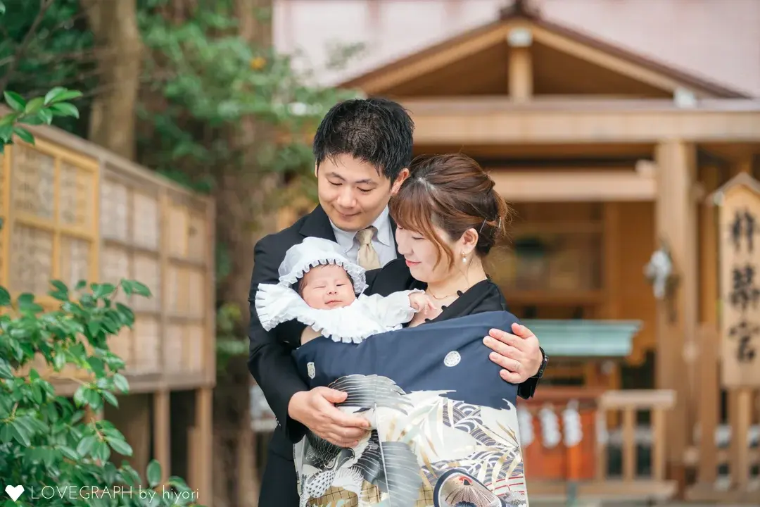 伊勢山皇大神宮の杵築宮・子之大神を背景に、お宮参りの記念撮影をする家族。スーツ姿のお母さんが白い帽子とドレスを着た赤ちゃんを抱き、背後からスーツ姿のお父さんが優しく手を添えている。赤ちゃんは鷹の柄が描かれた紺色の産着に包まれ、ご両親が愛おしそうにそのお顔を見つめている。