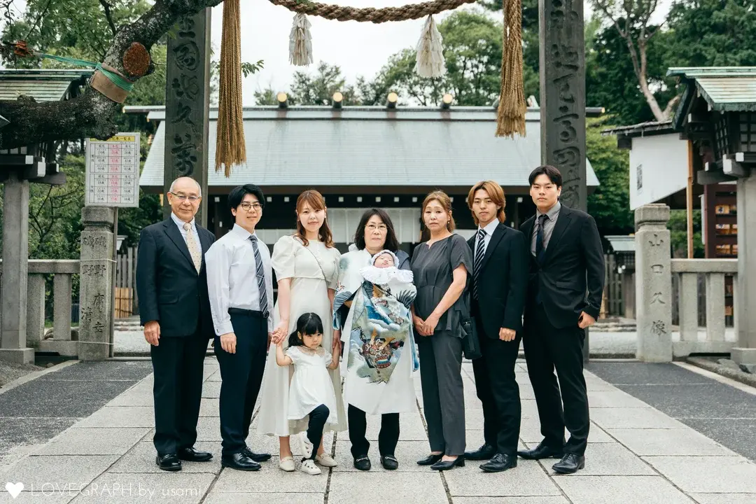 伊勢山皇大神宮の立派な鳥居と注連縄を背景に、横一列に並んでお宮参りの記念撮影をする8人のご家族。兜柄の産着を着た赤ちゃんを抱く女性を中心に、フォーマルな装いの大人たちと、お母さんと手を繋ぐ白いワンピース姿の小さな女の子が並んでいる。