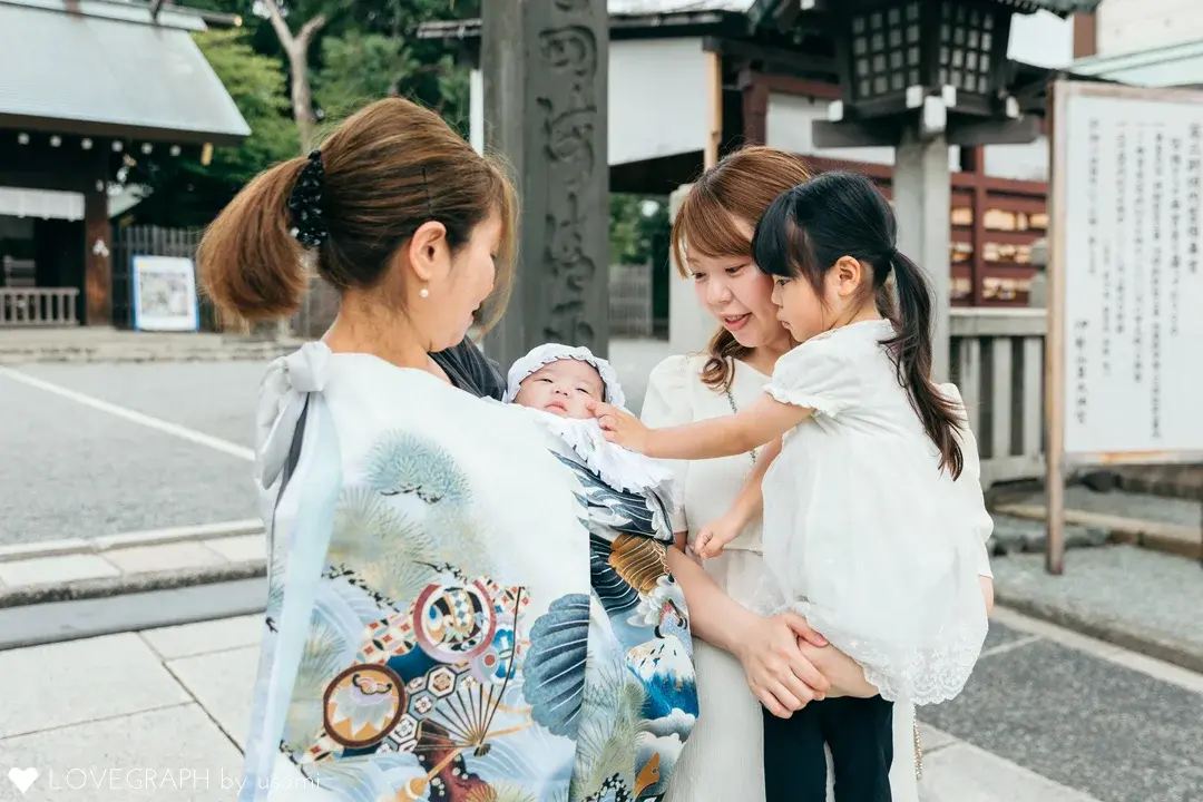 伊勢山皇大神宮の境内で、お宮参りのお祝いをするご家族。華やかな水色の産着に包まれた赤ちゃんを抱っこする女性と、白いワンピース姿の小さな女の子を抱っこして見守る女性が向かい合っている。女の子が赤ちゃんの頬に優しく手を伸ばしている心温まるシーン。