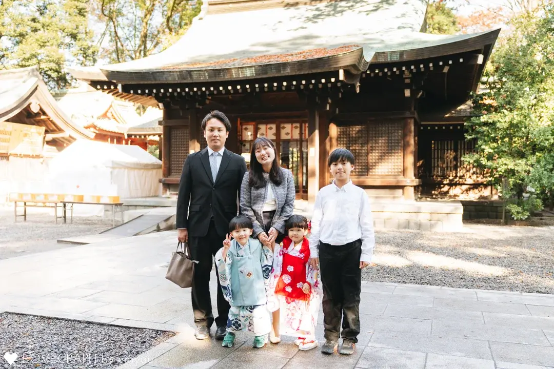 本殿・拝殿を背景に並ぶ5人家族の記念写真。手前には七五三を迎えた水色の袴の男の子と赤色の着物の女の子、後ろには両親とお兄ちゃんが並んで笑顔を見せている。