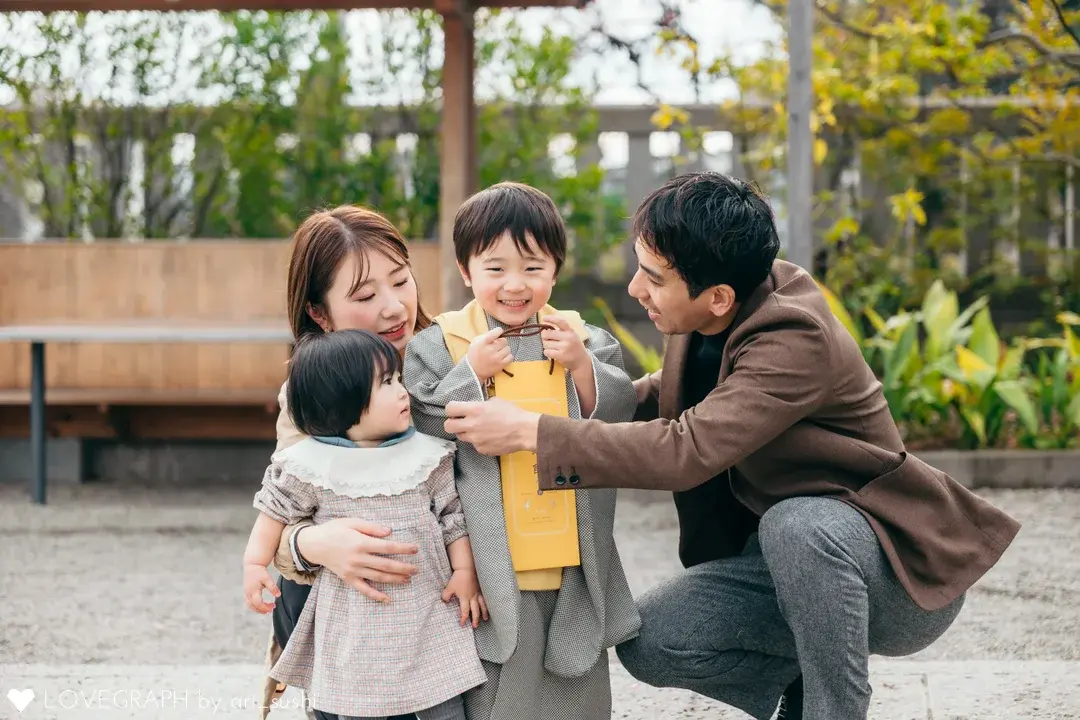 屋外で黄色の千歳飴の袋を胸に抱えて笑う袴姿の男の子と、その子に話しかける両親、母親に抱っこされた小さな妹の4人家族の自然な様子。