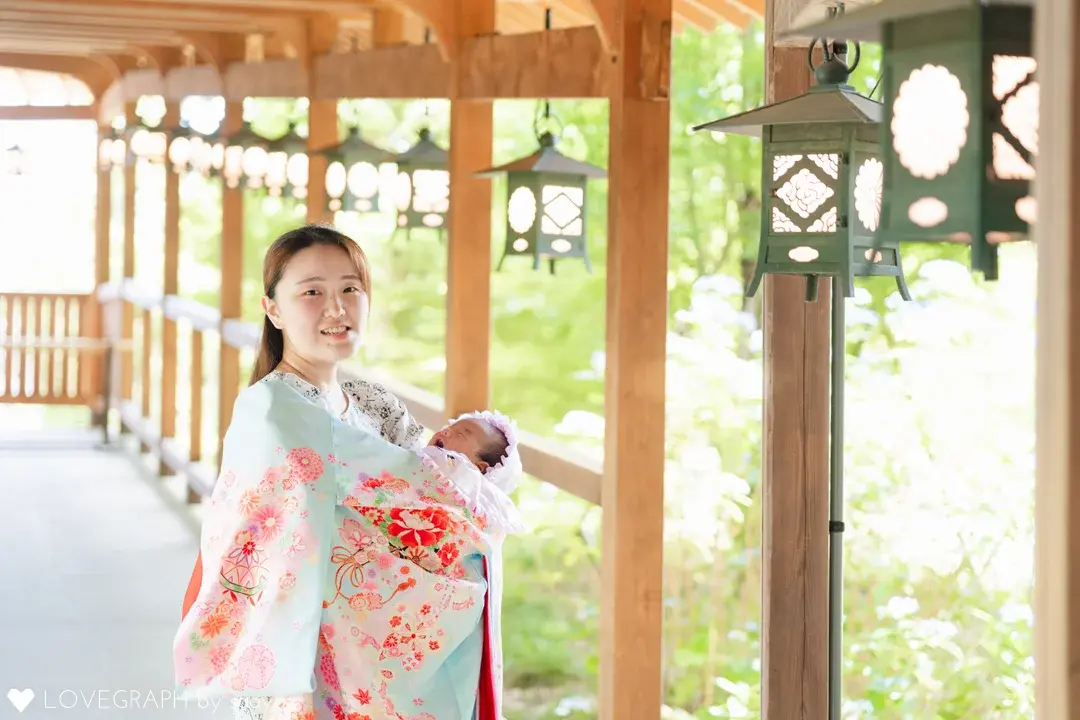 吊り灯籠が並ぶ川越氷川神社の回廊で、水色とピンクの花柄の産着に包まれた赤ちゃんを優しく抱っこしてカメラに微笑む母親のお宮参りの写真。背景には初夏の緑と紫陽花が見える。
