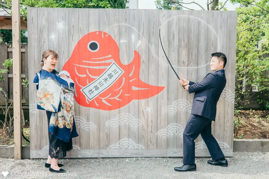 「川越氷川神社」と書かれた巨大な赤い鯛のイラストが描かれた板の前で、青い産着で赤ちゃんを抱っこして笑うママと、大きな釣竿の小道具を持って鯛を釣る真似をするパパのお宮参りの写真。
