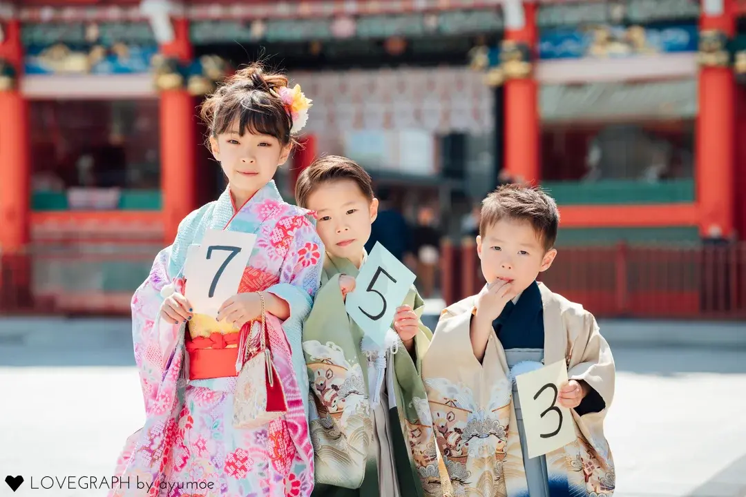 神田明神での七五三撮影にて、朱色の社殿を背に7歳・5歳・3歳の数字カードを持っておしゃれな着物で並ぶ3人きょうだいの出張撮影フォト